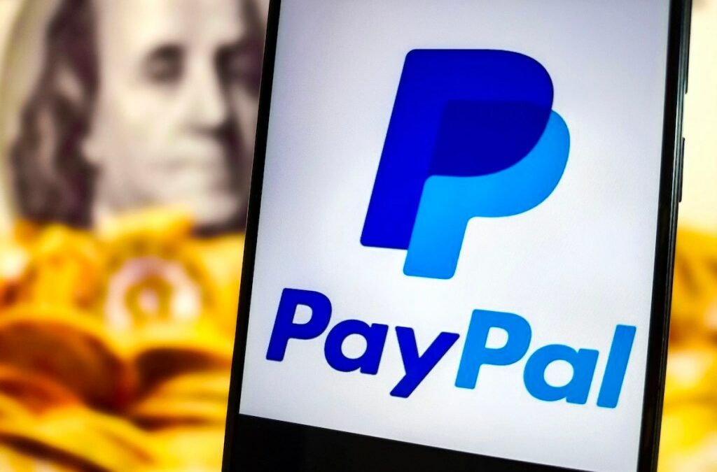 PayPal logo on a phone in front of a defocused background of gold coins and Benjamin Franklin's portrait from the US $100 bill