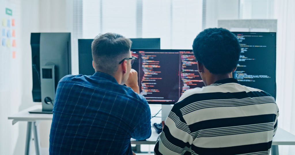 Two male software developers meeting on multiple computer screens displaying programming code, collaborating on project in office.